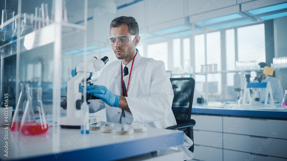 Medical Development Laboratory: Male Scientist is Using Microscope, Analyzes Petri Dish Sample. In Background Big Pharmaceutical Lab with Specialists Conducting Medicine, Biotechnology Research