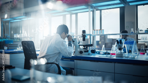 Medical Development Laboratory: Scientist Looking Under Microscope, Analyzes Petri Dish Sample. In Background Big Pharmaceutical Lab with Specialists Conducting Medicine, Biotechnology Research