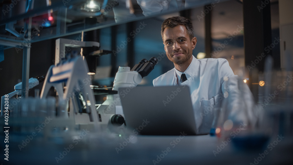 Medical Development Laboratory: Portrait of Male Scientist Works on Laptop, Looks at Camera and Smiles. Pharmaceutical Lab for Medicine, Biotechnology, Drugs, Research. Evening Work Medical Development Laboratory: Portrait of Male Scientist Works on Laptop, Looks at Camera and Smiles. Pharmaceutical Lab for Medicine, Biotechnology, Drugs, Research. Evening Work