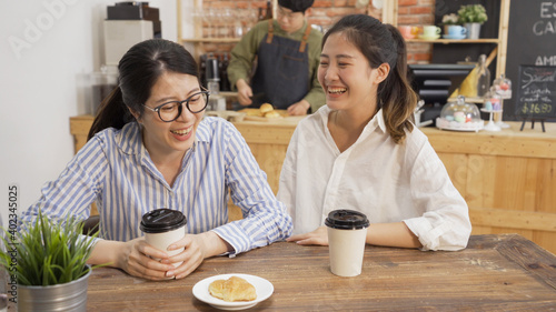 Two asian female coworkers talking at coffee shop while waiting for meal. girls colleagues friends chatting and laughing enjoy lunch break time together in cafe. man waiter prepare customer order
