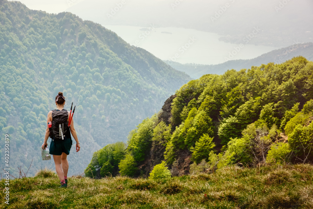 Girl hiker with backpack and trekking poles walk along a mountain path with a man on the hand in a sunny day. on the background the lake Maggiore, Val Grande National Park,  Europe, Piedmont Italy.