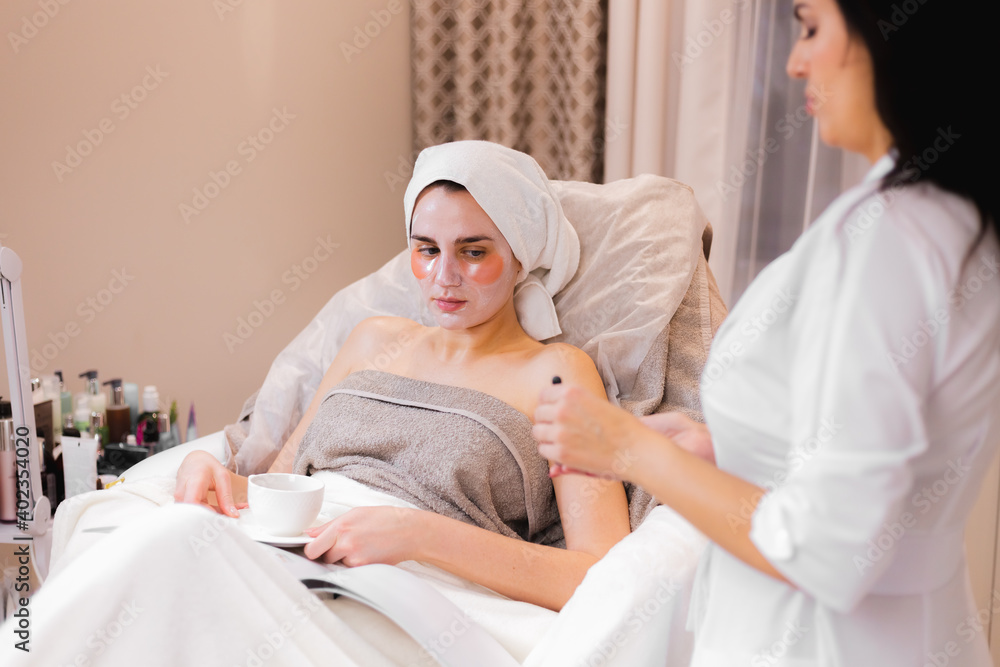A young girl in a beauty salon in a cosmetology office lies on the bed relaxes with a mask on her face and patches under her eyes, a beautician doctor stands next to A young girl in a beauty salon in a cosmetology office lies on the bed relaxes with a mask on her face and patches under her eyes, a beautician doctor stands next to