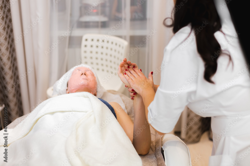 Obraz premium A young girl client with an alginate mask on her face lies on the cosmetology table, the beautician makes a manual hand massage