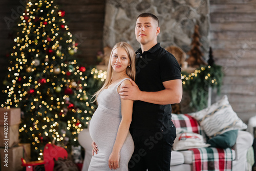 Adorable marriage couple near Christmas tree and fireplace at home, caring husband hug beautiful pregnant wife, young family enjoy happiness, winter holidays concept