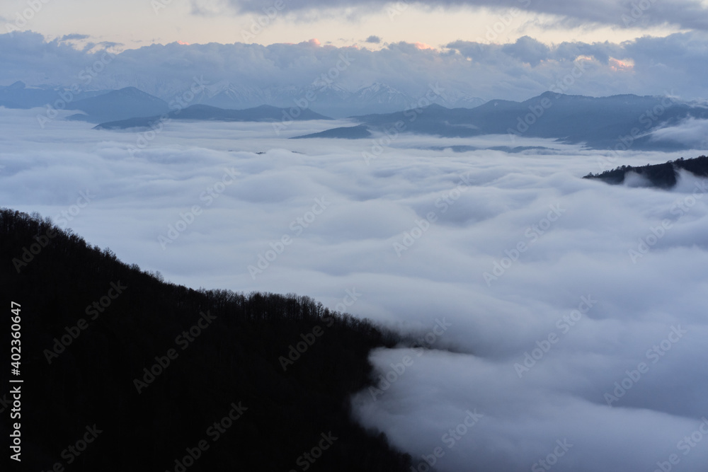 Low clouds, view of winter forest and mountains from observation deck, horizontal picture of amazing natural phenomenon. Beautiful unreal panoramic view of mountains in clouds.