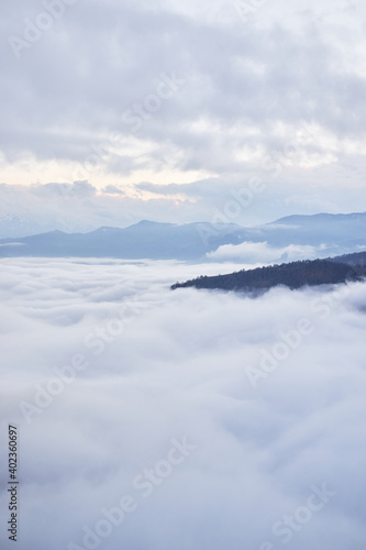 Low clouds, view of winter forest and mountains from observation deck, vertical picture of amazing natural phenomenon. Beautiful unreal panoramic view of mountains in clouds.