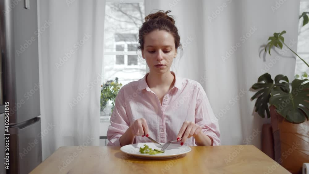 Young skinny woman sitting at table and eating piece of cucumber and salad with for and knife Young skinny woman sitting at table and eating piece of cucumber and salad with for and knife