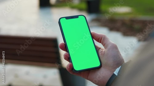 Outdoors young hands man holding use phone with vertical green screen in park background bench. Internet finger gadget device. Close up. Slow motion