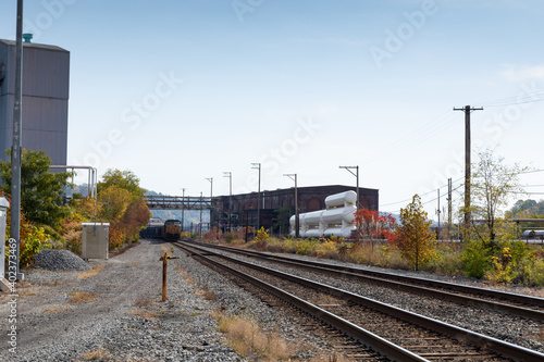 Train coming down a track running alongside industrial buildings in a rural area, fall season with autumn leaves, horizontal aspect