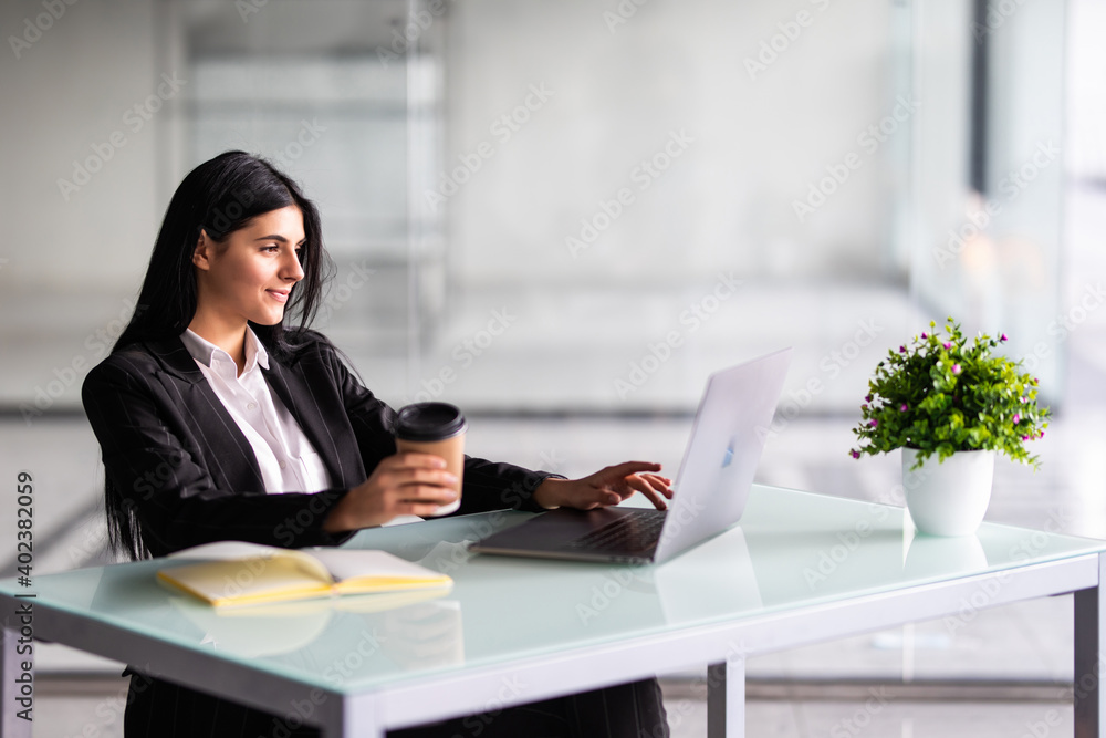Attractive woman sitting at desk in office, working with laptop holding document, having takeaway coffee. Attractive woman sitting at desk in office, working with laptop holding document, having takeaway coffee.