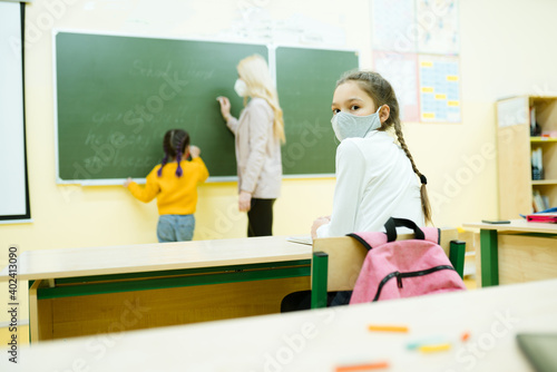 A student sits at a school desk while a teacher explains a lesson to a child in the background