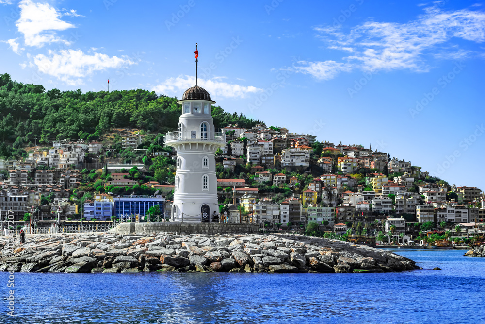 Alanya, Turkey - October 22,2020: Panorama of Alanya with a white old lighthouse in the foreground - view from the Mediterranean Sea. Seascape with cityscape on a hillside on the shore