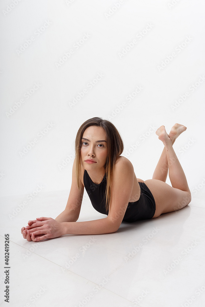 Obraz premium sexy asian woman with long hair posing in black lingerie on white studio background with bare feet. attractive female lying on floor on her belly. model tests of skinny lady in bodysuit