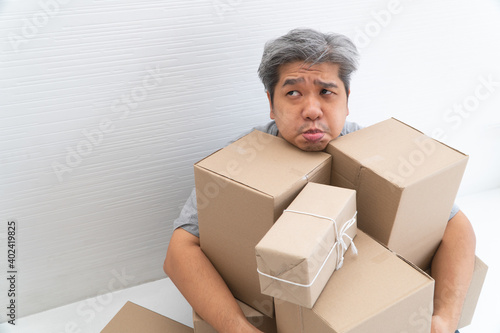 Asian Shopaholic man Shocked and sitting on the floor in the living room and Cardboard Box on top of him after The courier delivered to the home. Concept of shopping online and Shopaholic.