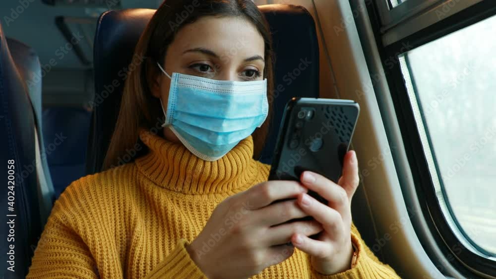 Young woman in protective medical face mask messaging with smartphone on train Young woman in protective medical face mask messaging with smartphone on train