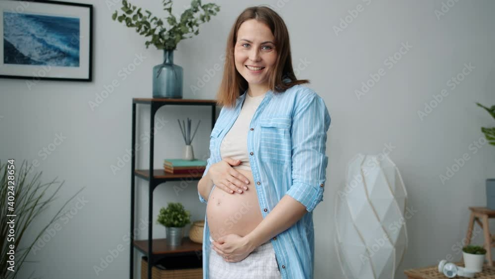Slow motion portrait of cheerful pregnant lady touching belly and looking at camera smiling feeling love towards baby standing alone at home. Pregnancy and emotions concept. Slow motion portrait of cheerful pregnant lady touching belly and looking at camera smiling feeling love towards baby standing alone at home. Pregnancy and emotions concept.
