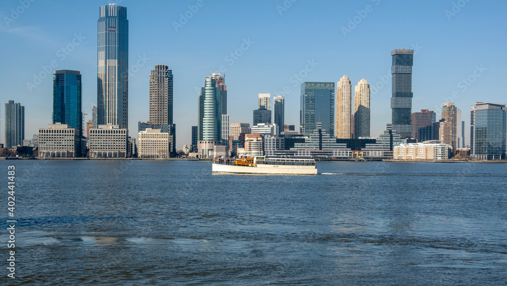 Obraz premium New York, USA, December 2020. A tourist boat on the River Hudson with a backdrop of the Colgate Clock and Jersey City highrise buildings, during the Covid pandemic, from Downtown Manhattan.