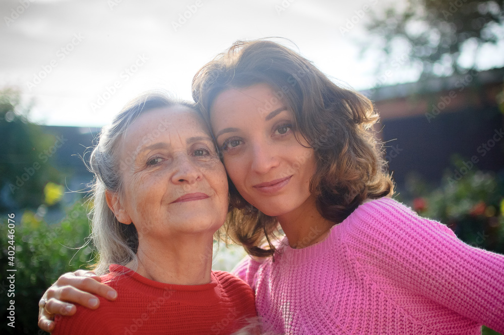 Senior mother with gray hair with her adult daughter looking at the camera in the garden and hugging each other during sunny day outdoors Senior mother with gray hair with her adult daughter looking at the camera in the garden and hugging each other during sunny day outdoors