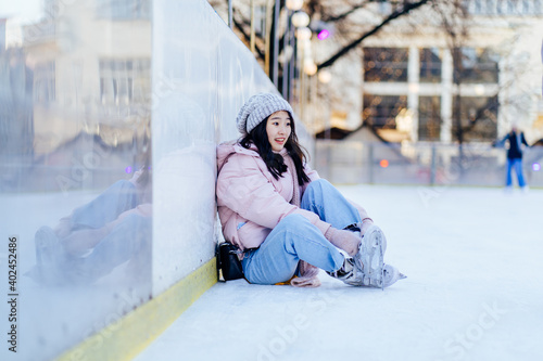 Ice skating concept. Surprise emotion asian woman sitting on the ice rink. Fall on ice. Korean girl tying shoelaces on ice skates fell on ice in european city square, outdoor. Winter sunny frozy day. photo