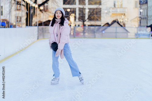 Full height of millennial of a young asian female wearing powder pink jacket and warm knitted hat, scarf and gloves skating on ice rink, outdoors at winter time. © Iryna