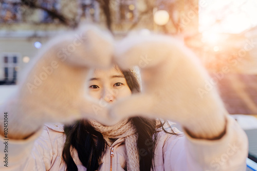 Lovely korean female doing heart shape with hand and fingers in fluffy knitted gloves smiling looking through sign while to skate on the rink cheerful and excited outdoors.