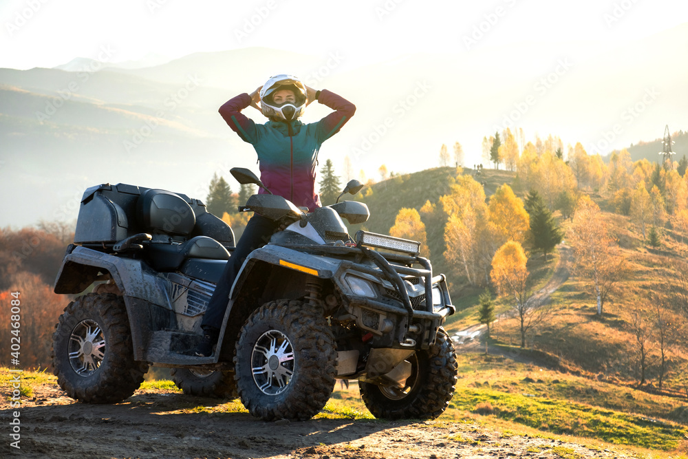 Happy active female driver in protective helmet enjoying extreme riding on ATV quad motorbike in fall mountains at sunset. - obrazy, fototapety, plakaty 