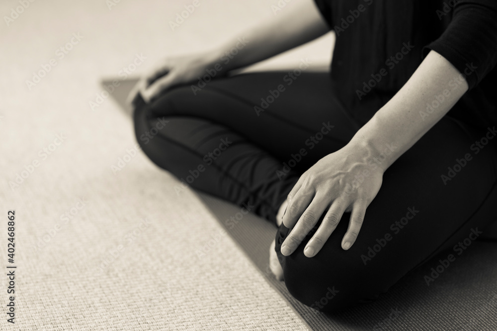 Horizontal side closeup of lower body of yogini on the floor with legs crossed in lotus pose. Woman indoors wearing black yoga pants with hands resting on knees. Home yoga practice in sepia tone