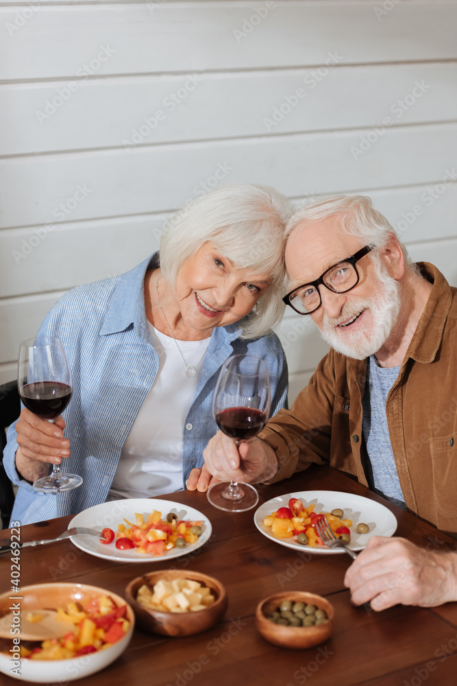 happy senior couple with wine glasses looking at camera while sitting at table with vegetarian dinner on blurred foreground happy senior couple with wine glasses looking at camera while sitting at table with vegetarian dinner on blurred foreground