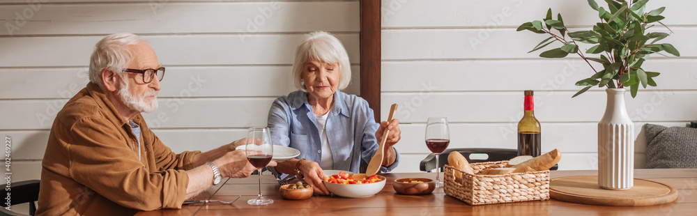 smiling senior wife with spatula serving salad for husband at table with vegetarian dinner in kitchen, banner smiling senior wife with spatula serving salad for husband at table with vegetarian dinner in kitchen, banner
