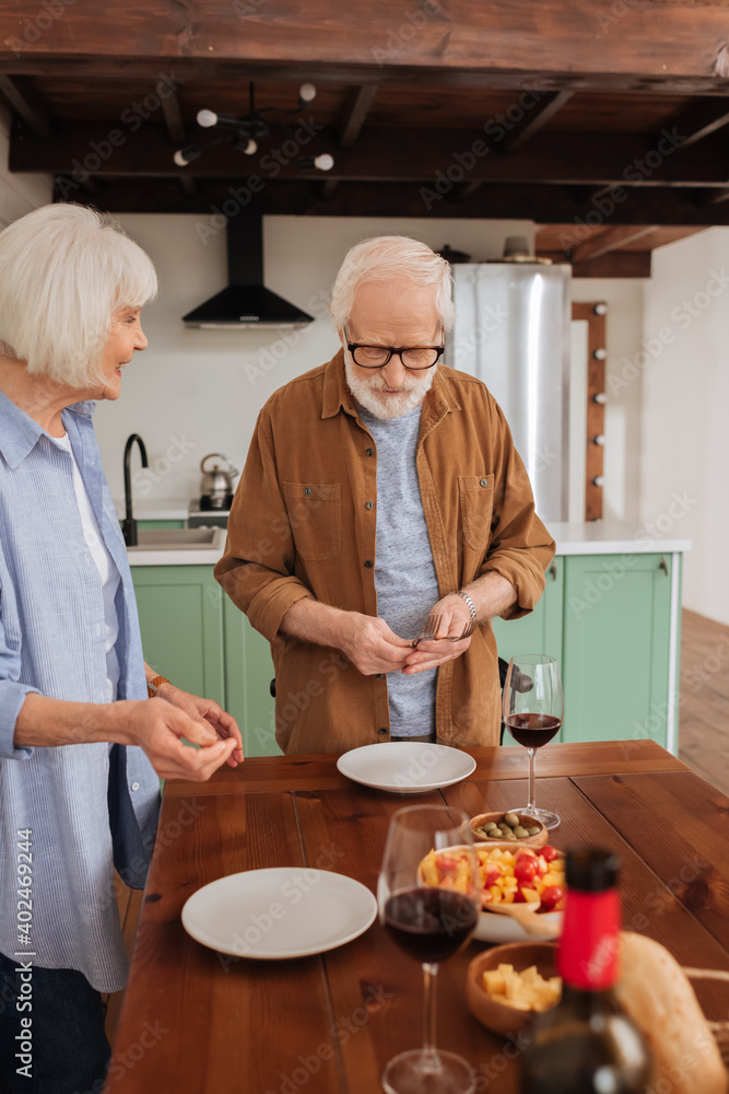 smiling senior wife looking at husband serving table with forks in kitchen on blurred foreground smiling senior wife looking at husband serving table with forks in kitchen on blurred foreground