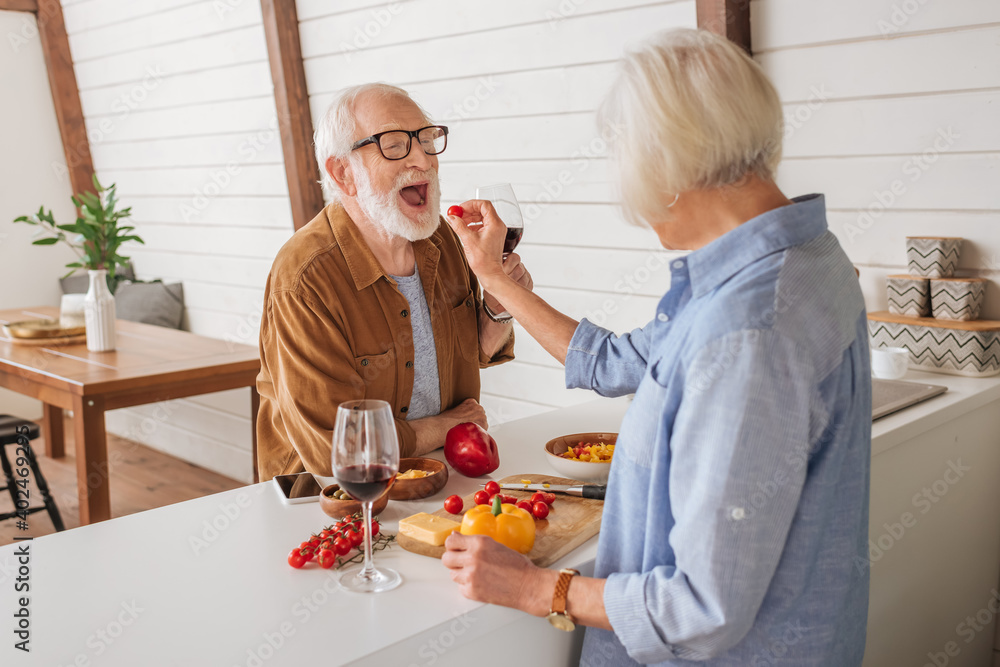 back view of elderly wife feeding happy husband with cherry tomato near table with food in kitchen back view of elderly wife feeding happy husband with cherry tomato near table with food in kitchen
