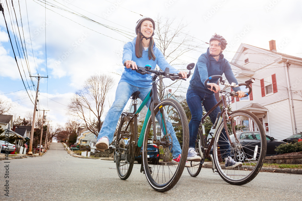 Low angle portrait of a laughing couple with smiling young woman ride a bicycle on the urban street wearing helmets