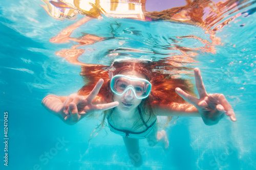 Smiling happy portrait in scuba mask of the girl swim underwater in the pool and showing v victory gesture with hands