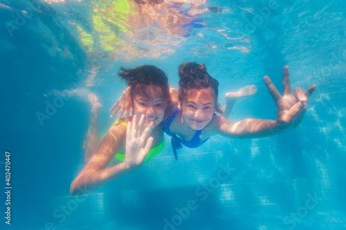 Two happy teenage girls swim together hugging underwater in the pool waving hands