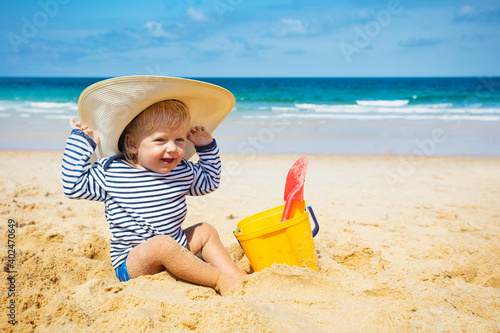 Happy little toddler boy sit on the beach wear big straw hat play with plastic bucket