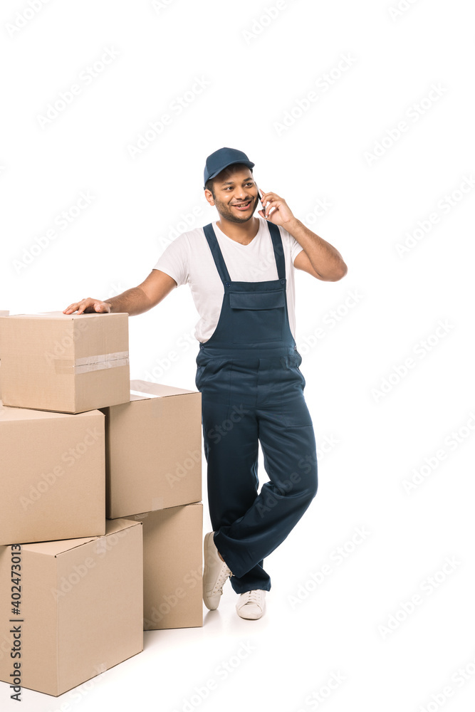 full length of happy indian mover in overalls talking on smartphone while standing near carton boxes on white full length of happy indian mover in overalls talking on smartphone while standing near carton boxes on white