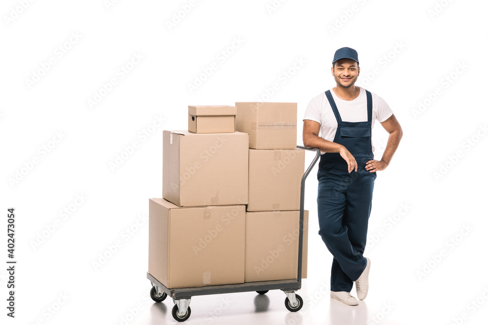 full length of cheerful indian mover in overalls standing with hand on hip near hand truck with carton boxes on white full length of cheerful indian mover in overalls standing with hand on hip near hand truck with carton boxes on white