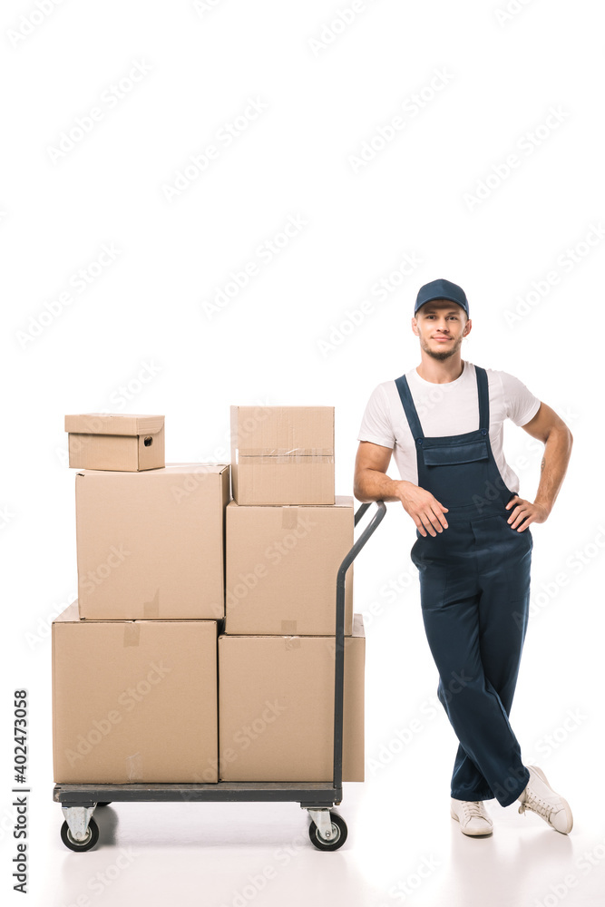 full length of cheerful mover in uniform and cap leaning on hand truck with carton boxes on white full length of cheerful mover in uniform and cap leaning on hand truck with carton boxes on white