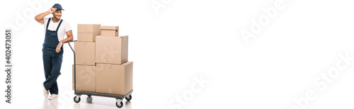 full length of joyful indian mover standing near hand truck with carton boxes and adjusting cap on white, banner © LIGHTFIELD STUDIOS