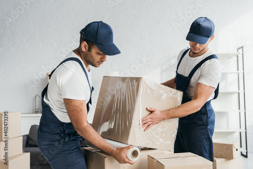 Quadro su tela multiethnic movers in overalls wrapping box with stretch film