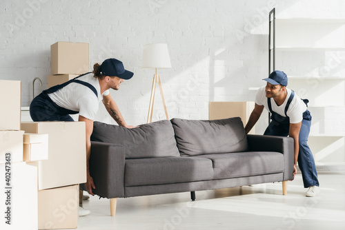 happy multicultural movers in uniform smiling while carrying couch in apartment photo