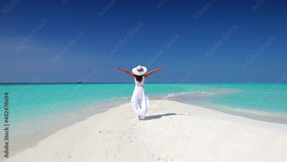 A woman with white hat and outstretched arms walks over a tropical beach towards turquoise ocean and enjoys her exotic getaway