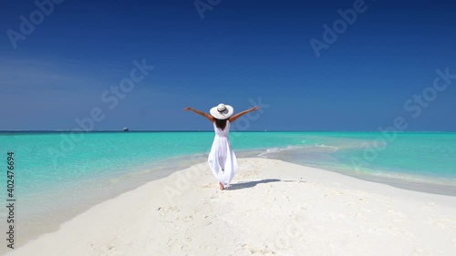 A woman with white hat and outstretched arms walks over a tropical beach towards turquoise ocean and enjoys her exotic getaway