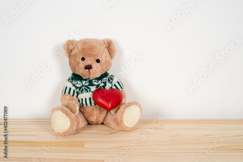 One teddy bear carrying a red wooden heart on his white and dark green pullover, sitting on a wooden table and leaning against a white wall as background 