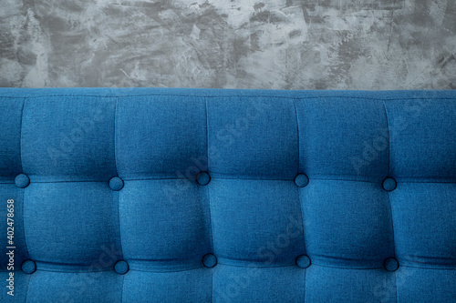 A piece of upholstered furniture with classic Chesterfield pattern with buttons on grey concrete wall background. Modern blue furniture fabric. Elegant luxury sofa upholstery. Interior backdrop