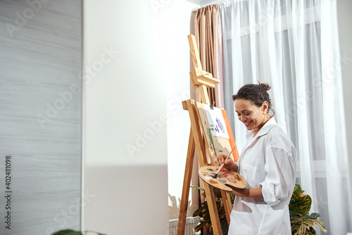 View of a smiling female painter with gathered hair in a bun and paint brushes in her hair standing in front of the easel in workshop and drawing