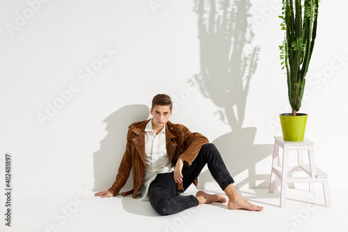 handsome man in brown jacket and trousers sits on the floor near a flower in a pot