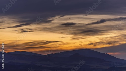 Timelapse of a sunset setting over a mountainous landscape in the sky with colorful moving clouds of orange color and fog moving on a hill.