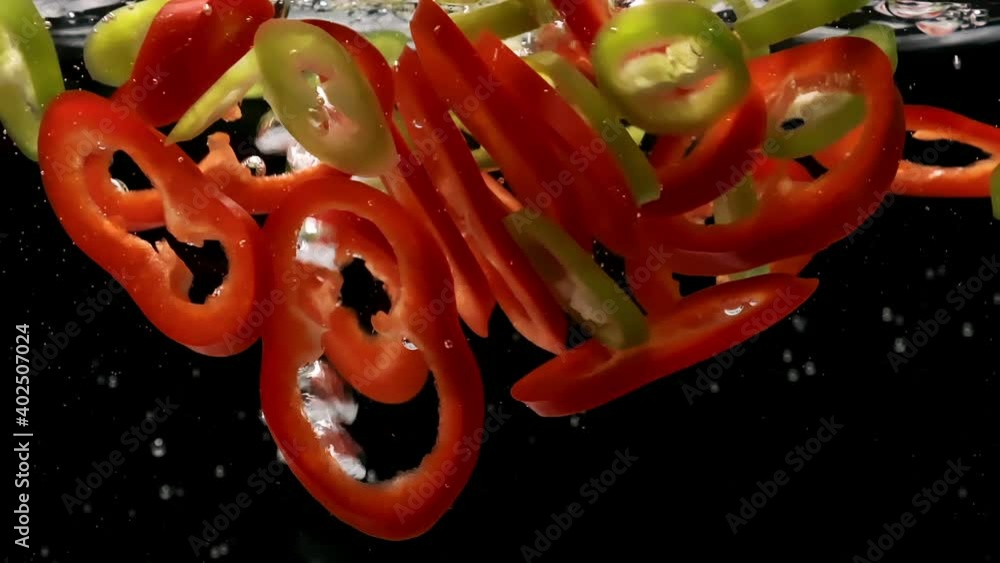 Red and green ripe peppers slices fall down into water and bubbles rise upward on black background extreme closeup slow motion Red and green ripe peppers slices fall down into water and bubbles rise upward on black background extreme closeup slow motion