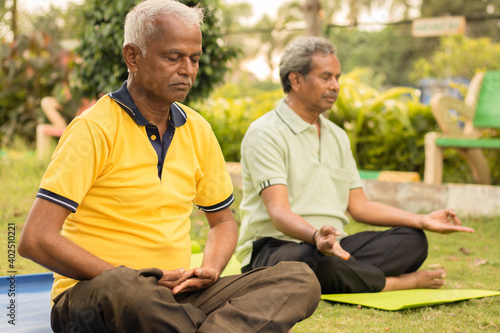 Two Indian senior men doing meditation on yoga mat at park by closing eyes - concept of elderly healthcare and lifestyle